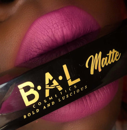 A model holds the Bold and Luscious Brand Lip stick bottle in between her lips wearing the color on her lips. Her lips are  fuchsia matte and the color is not overly bright. 