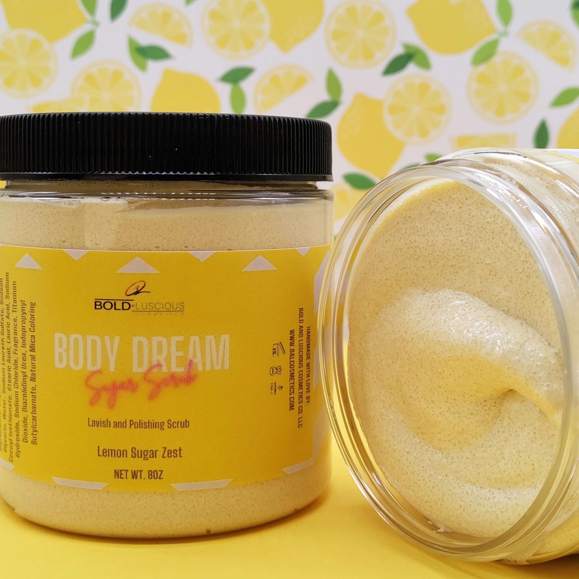 There are two Bold and Luscious Cosmetics Lemon Zest scrubs in this picture for illustration. One is the closed bottle of 8 ounces of scrub with a yellow label. The second is an open container showing its thick and creamy texture.  The background style has a pattern of lemons to give this image a vibrant feel.