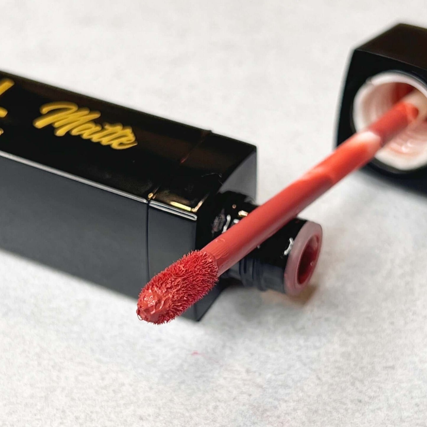 From the left you see a blurred picture of the right side of the bottle of Bold and Luscious Lipstick. The tip of the bottle is fluffy and you can see a muted rose color on the edge of the soft brush.