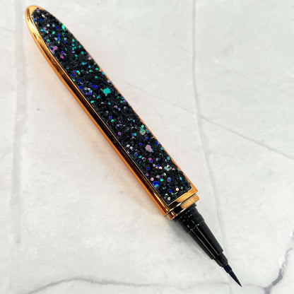 Bold and Luscious Diamond Status Magic Lash Adhesive Liner is a flat and wide style pen casing with a fine tip for precision use. a full shot reveals the beautiful casing with blue, black purple and turquoise glitter on the outside and a gold trim.