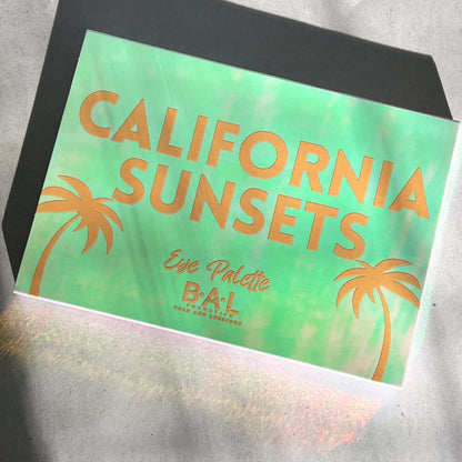 A Closed Eye Shadow Palette lays in the sunlight. It reads in yellow leads California Sunsets Palette by Bold and Luscious Cosmetics.