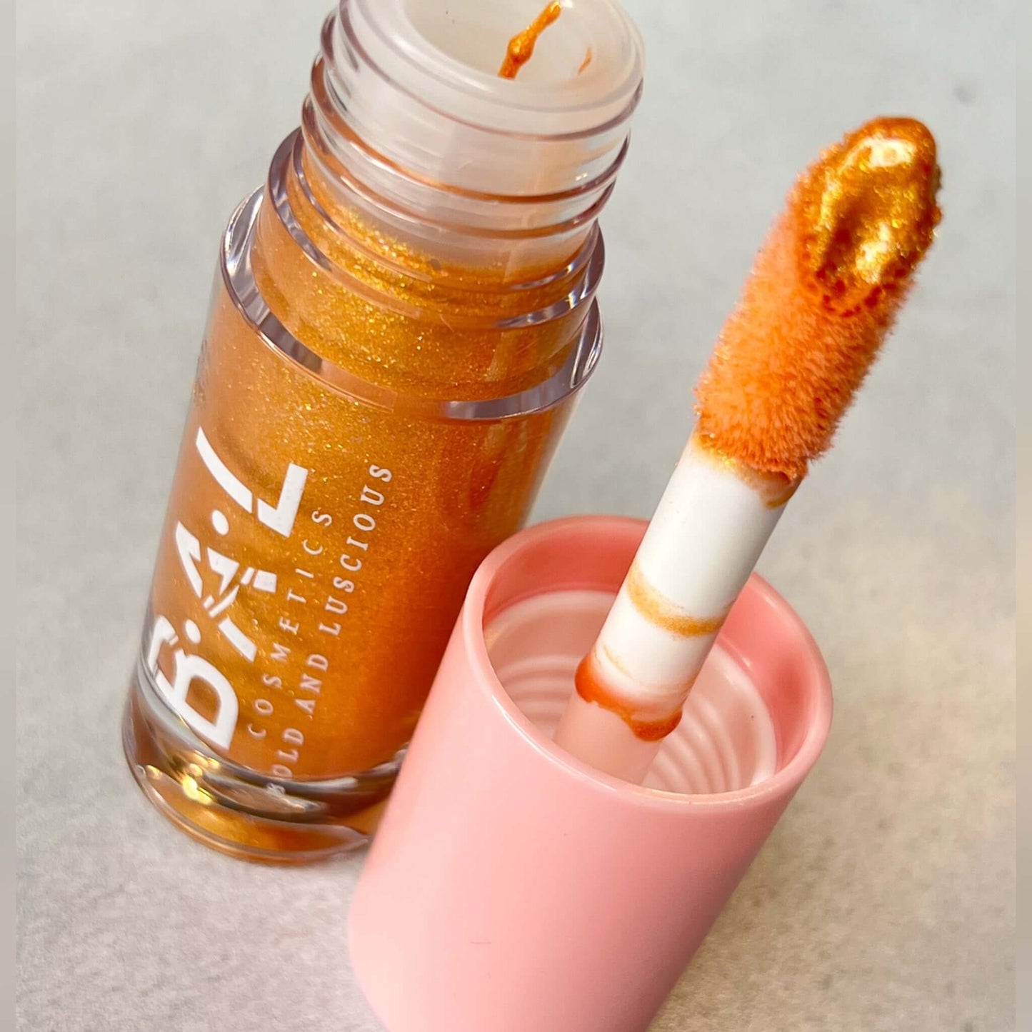 Bold and Luscious Cosmetics Lip Gloss in color Champagne. In the top left is the bottle of the gloss without the lid. the applicator is standing on its top next to the gloss. You can see the golden color glitter gloss and its rich color. 