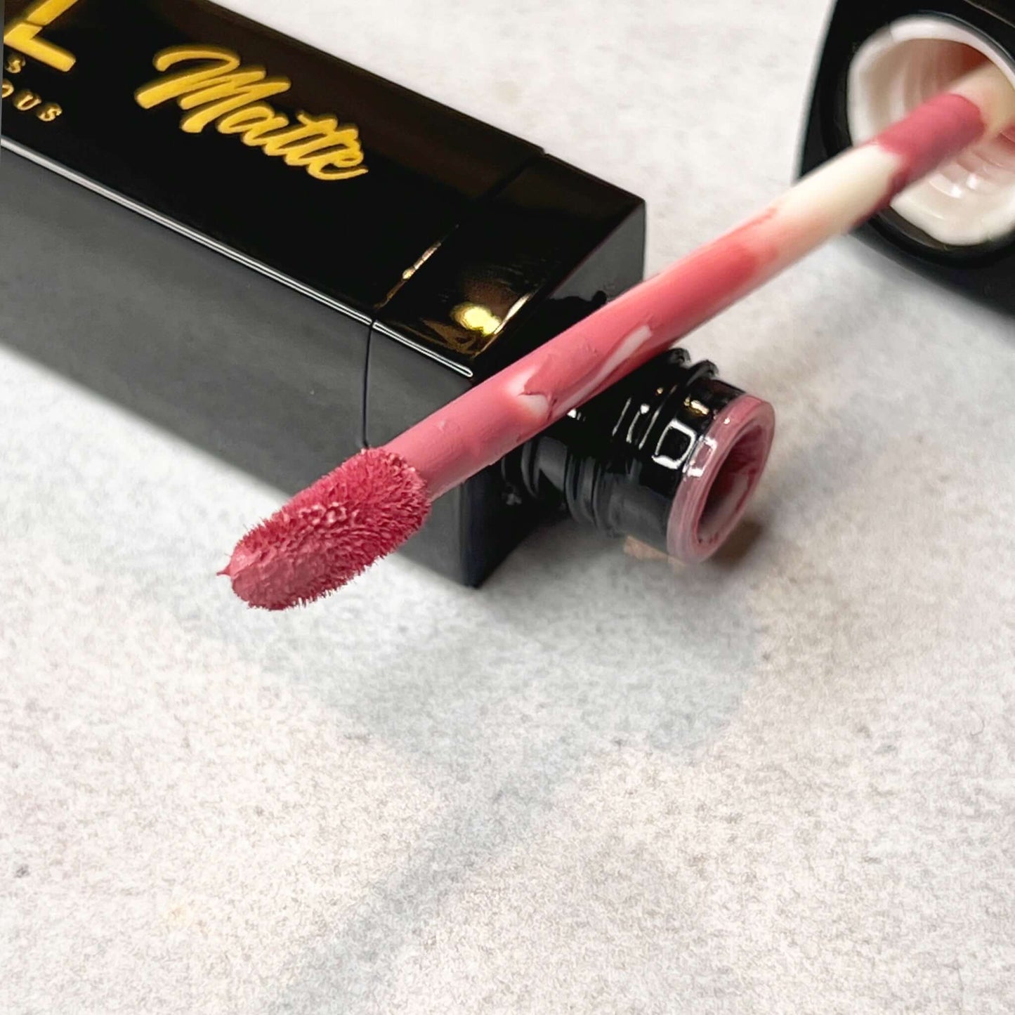 A close up of the Lipstick wand reveals a muted frosted pink color on the tip of a soft fluffy brush resting against a bold and luscious cosmetics matte lipstick bottle